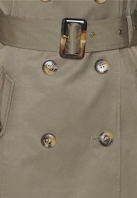 Khaki płaszcz trench z podwójnym zapięciem na guziki, dużymi guzikami z detalem logo, teksturowanym materiałem oraz dopasowanym paskiem z prostokątną klamrą.