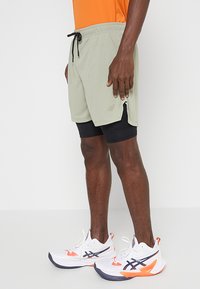 Pantalones cortos deportivos verde claro con cintura elástica y cordón, combinados con pantalones cortos de compresión negros debajo. Zapatos deportivos blancos y naranjas visibles.