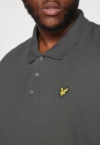 Ingrandimento di una persona che indossa una polo grigia scura con tre bottoni e un piccolo logo di uccellino giallo sul petto.