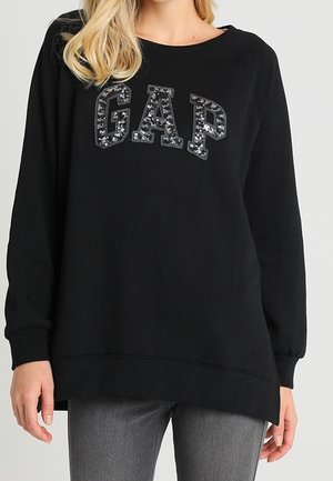 Kvinde iført en sort sweatshirt med lange ærmer og et glitrende "GAP" logo, sammen med grå jeans, foran en ensfarvet baggrund.