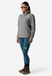 Pullover in pile grigio con zip a metà, leggings turchesi e scarponi da trekking marroni. Presenta loghi bianchi e una vestibilità comoda.