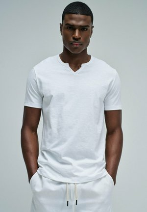 Joven con cabello corto que lleva una camiseta blanca lisa con cuello en V y pantalones blancos, con las manos en los bolsillos, fondo gris neutro.