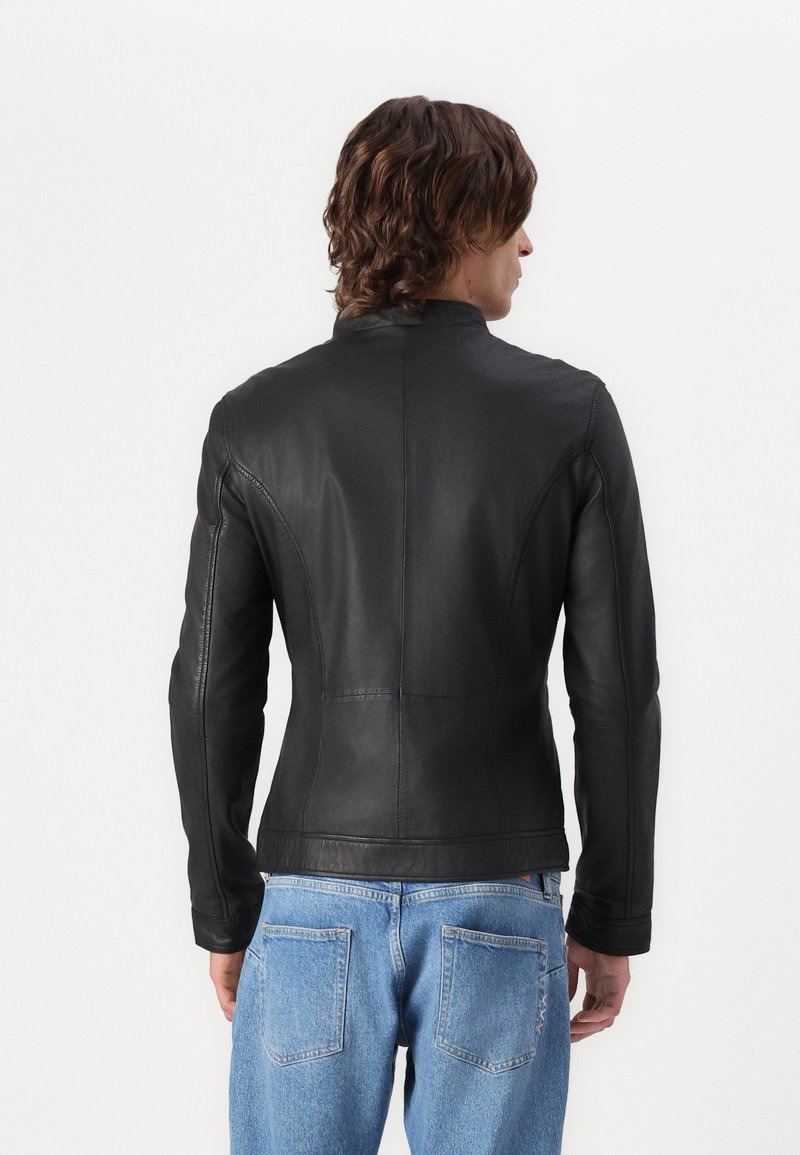 Schwarze Lederjacke mit taillierter Passform, hohem Kragen und nahtloser Verarbeitung; getragen über hellblauen Jeans. Die Textur wirkt glatt und poliert.