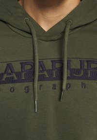 Oliwkowy hoodie z kieszenią kangurką, kapturem na sznurku i haftowanym granatowym logo na klatce piersiowej. Gładka tekstura.