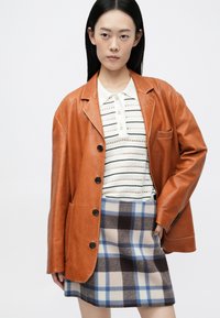 Brun læderblazer med en afslappet pasform, sorte knapper, parret med en stribet polo t-shirt og en rutet mini-nederdel i blå og beige nuancer.