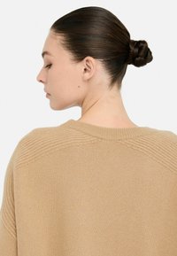 Maglione beige in maglia con cuciture a coste sulle spalle, che presenta un colletto rotondo e una texture morbida. Lo sfondo è bianco semplice.