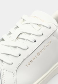 Baskets blanches avec un dessus en cuir lisse, bout arrondi, lacets plats et logo doré "TOMMY HILFIGER" sur le côté.