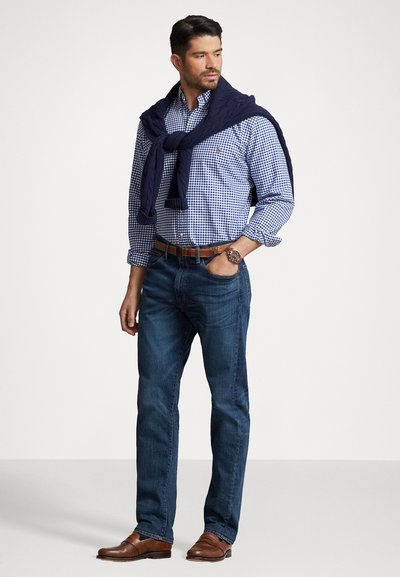 Kollande skjorta i blått rutmönster, marinblå tröja slingrande över axlarna, blå jeans och bruna läderskor med detaljerad sömnad.
