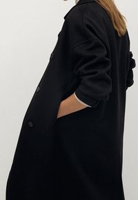 Abrigo de lana negro con un corte holgado, grandes botones y mangas remangadas, con bolsillos laterales y un detalle en el collar.