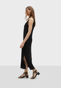 Crna haljina bez rukava s vertikalnim nabora, bočnim prorezima, uparenih s crnim sandalama s trakama koje imaju zlatne studove i blok potpetice.