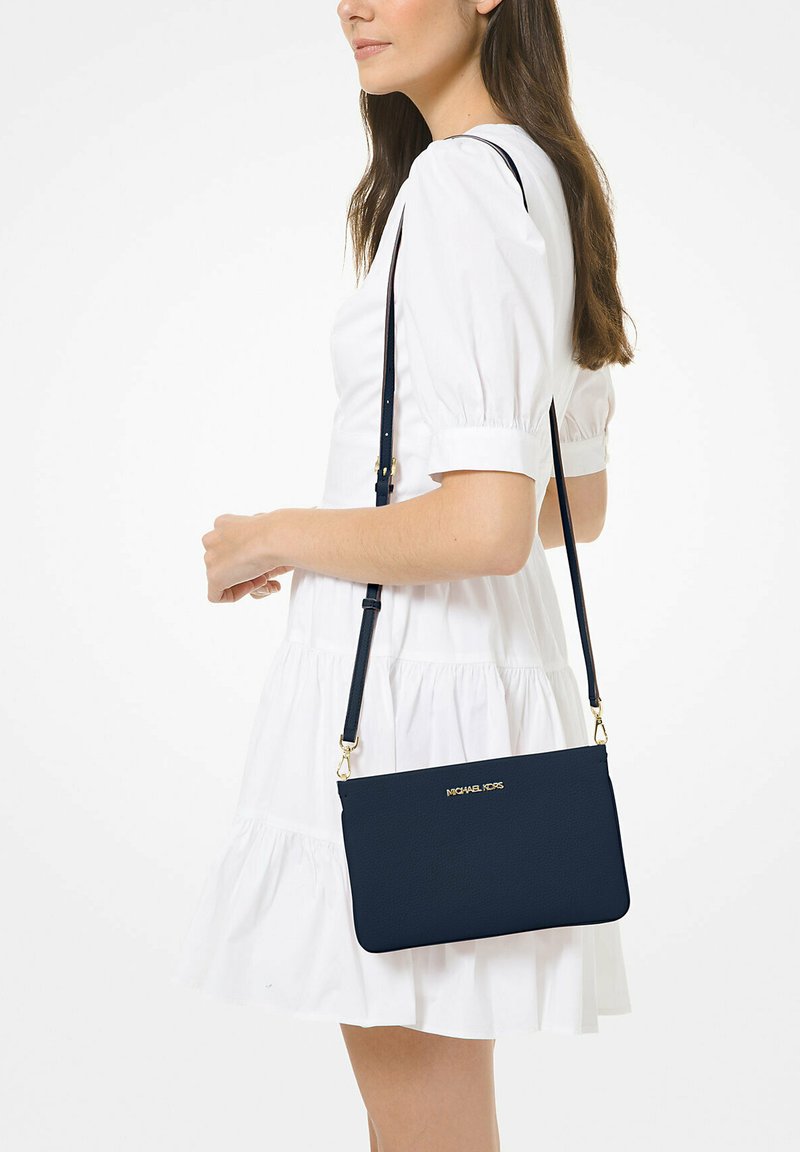 Borsa a tracolla blu navy con finitura testurizzata, dettagli della logo in oro e tracolla regolabile, indossata sopra un abito bianco da un modello.