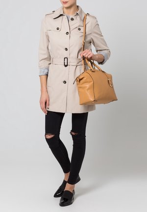 Frau trägt beige trenchcoat mit Gürtel, schwarze zerrissene Skinny-Jeans, schwarze Loafer und eine tanfarbene Ledertasche über der Schulter.