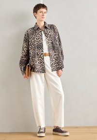 Personne debout portant une chemise à imprimé léopard, un débardeur blanc, un pantalon blanc, des chaussures à imprimé léopard, tenant une pochette couleur sable sur un fond uni.