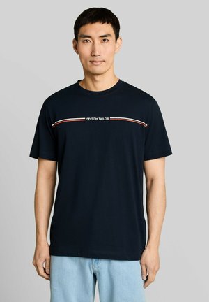 Mann trägt ein schwarzes Tom Tailor T-Shirt mit roten und weißen Streifen quer über der Brust und hellblaue Jeans, steht vor einem grauen Hintergrund.