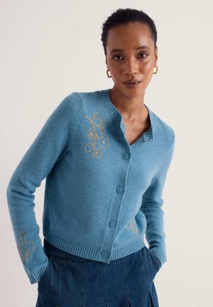 Femme portant un cardigan bleu à boutons avec une broderie florale jaune, jupe bleu foncé, boucles d'oreilles créoles dorées, mains dans les poches, fond neutre.