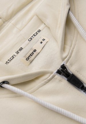 Close-up of beige hoodie neckline showing two fabric labels reading "ombre" and a black zipper pull with "ombre" text.