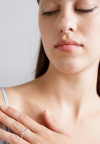 Mladá žena so zatvorenými očami sa dotýka svojho ľavého ramena, na ľavej ruke má na prsteni dekoratívny prsteň.