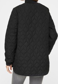 Person som bär en svart vadderad jacka med diamantmönster, ribbade ärmar och ljusgrå byxor, sedd från baksidan.