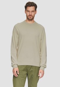 Felpa beige con maniche raglan, colletto a girocollo e polsini a coste. Texture morbida con minimo dettaglio di cucitura. Abbinata a pantaloni verdi.