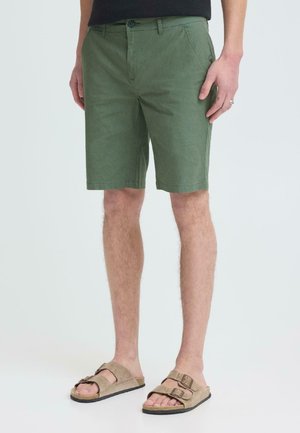 Man die olijfgroene knielange korte broek en beige sandalen met gesp draagt, staand tegen een effen lichte achtergrond.