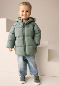Young child wearing a green hooded puffer jacket, loose blue jeans with small rips, and green sneakers, standing and smiling indoors.