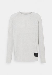 Chemise à manches longues gris clair en tissu doux et texturé. Comprend des manches raglan et un patch accent noir sur le côté inférieur gauche.