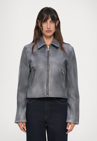 Mujer que lleva una chaqueta de cuero gris con cremallera y bolsillos laterales, y jeans oscuros de cintura alta, de pie contra un fondo blanco liso.