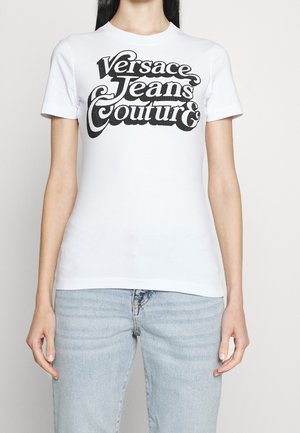 Kobieta w białej koszulce z krótkim rękawem z czarnym tekstem "Versace Jeans Couture" i jasnoniebieskich dżinsach na jednolitym tle.