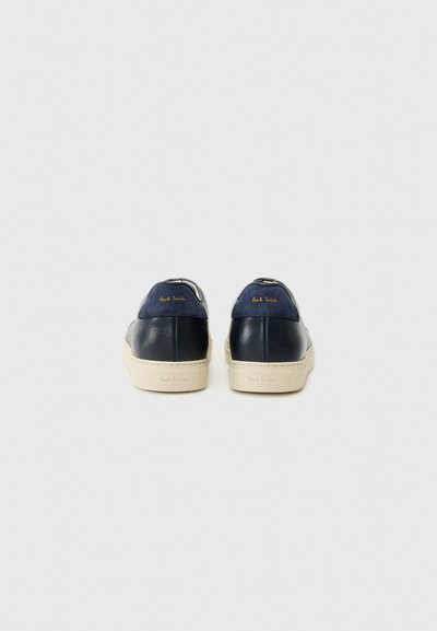 Sapatilhas de pele azul-marinho com uma sola de borracha creme; apresentam uma textura suave e detalhe em logótipo dourado no calcanhar. Design minimalista.