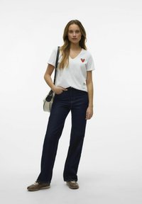 T-shirt blanc à col en V avec un motif de cœur rouge et vert sur la poitrine, associé à un jean large bleu foncé et des chaussures marron.