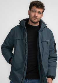 Tyrkis isoleret jakke med lynlås i front, hætte og flere lommer. Glat tekstur og semiløs pasform, parret med en sort, ribbet sweater under.