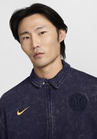 Nuori mies, jolla on mustat hiukset, pukeutuneena tummansiniseen Nike-vetoketjulliseen takkiin, jossa on hienovarainen logo ja keltainen Nike-swoosh yksinkertaisella taustalla.