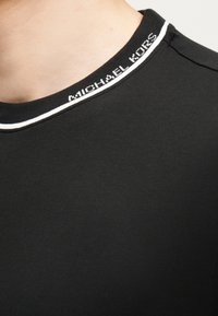 Κοντινή φωτογραφία του γιακά μαύρου μπλουζιού με στρογγυλή λαιμόκοψη, επάνω στην οποία υπάρχει λευκή λεπτομέρεια και το κείμενο "MICHAEL KORS" υφασμένο στη λαιμόκοψη.