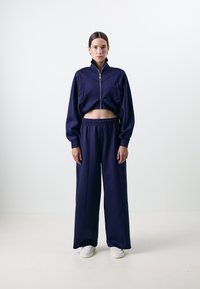 Casaco curto azul marinho com fecho e mangas compridas, combinado com calças largas também em azul marinho. Ambos feitos de um tecido liso e leve. Ténis brancos.