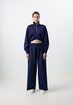 Marineblå cropped jakke med glidelås og lange ermer, kombinert med vide marineblå bukser. Begge laget av glatt, lett stoff. Hvide sneakers.