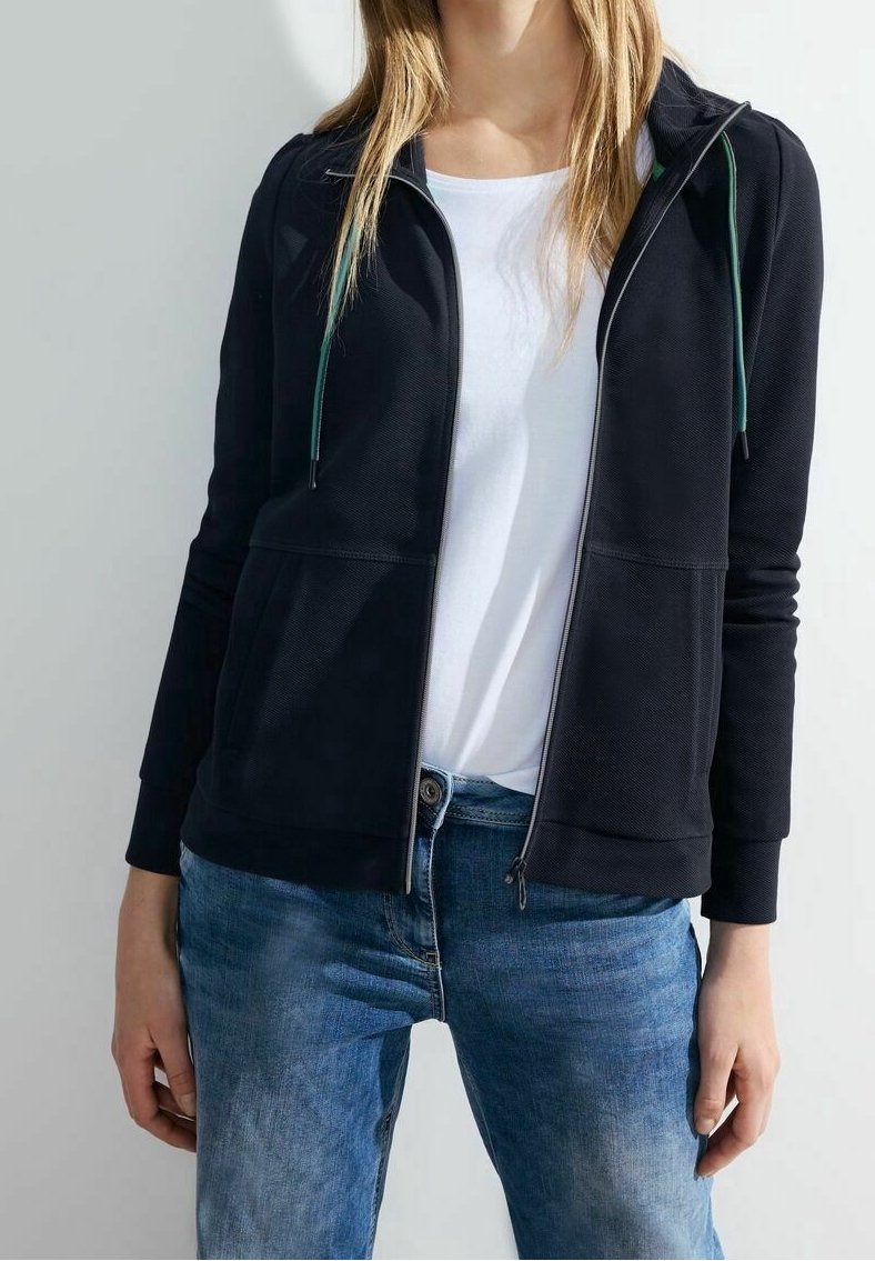 Personne portant un sweat à capuche zippé noir avec des cordons verts, un t-shirt blanc et un jean bleu, montrée du cou aux hanches.