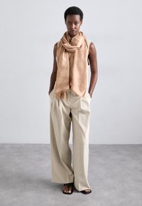 Bufanda beige con un patrón sutil colocada alrededor del cuello, combinada con pantalones sueltos de un gris claro y sandalias planas negras.