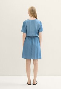 Vrouw met lang blond haar, gekleed in een blauw jurkje met korte mouwen tot op de knie en zwarte instap sandalen, staat met haar rug naar de kijker.