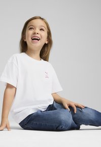 Hvid bomulds t-shirt med et pink Puma-logo på venstre bryst, parret med blå denim jeans, siddende på en lys grå overflade.