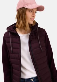 Femme portant une casquette rose, une veste matelassée bordeaux avec capuche, une chemise blanche et un jean bleu, regardant sur le côté sur un fond blanc.