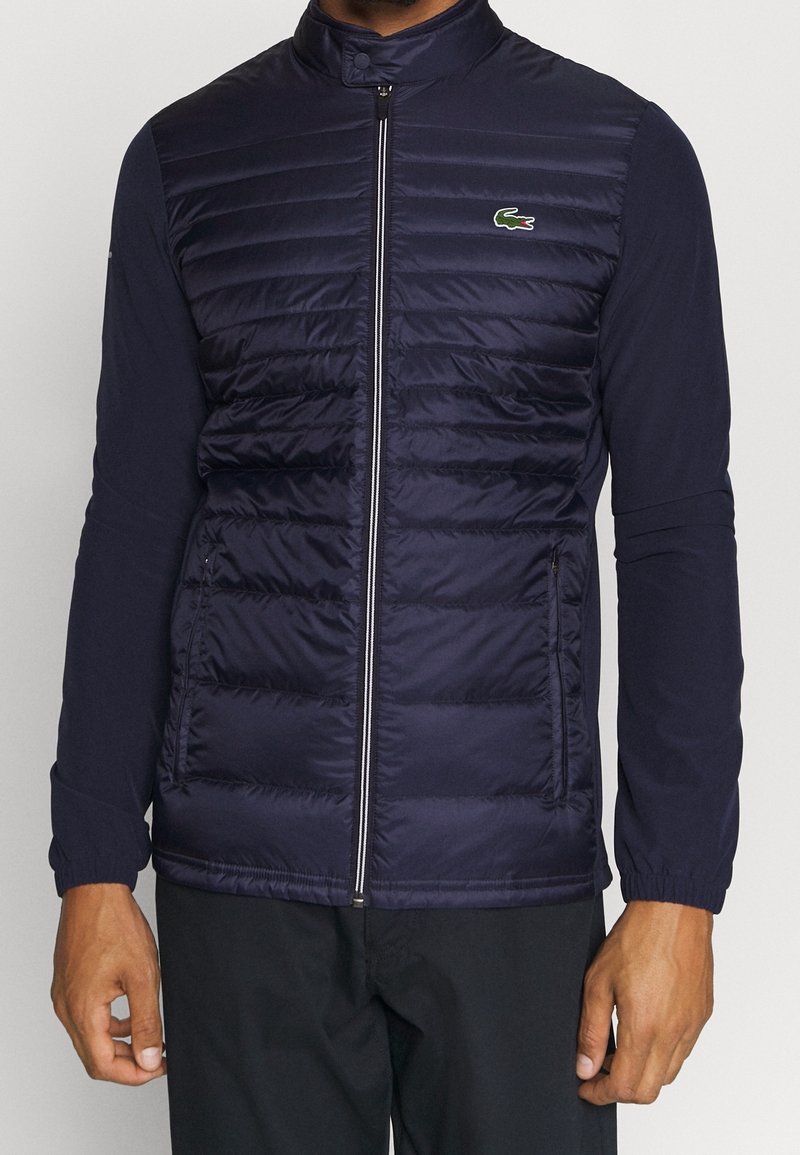 Marinblå pufferjacka med hög krage, blixtlås framtill och texturerade paneler. Har långa ärmar med elastiska manschetter och en grön logotyp på bröstet.