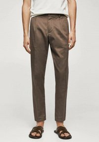 Pantalones marrones ajustados con una textura suave, pliegues frontales, bolsillos laterales y un diseño de pierna recta. Combinados con sandalias negras.