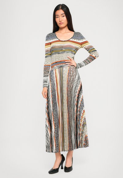 Donna che indossa un vestito midi a maniche lunghe, a strisce e motivi multicolori, con tacchi alti neri, in piedi con una mano sui fianchi.
