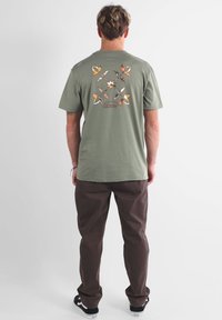 T-shirt vert en coton avec un graphique de papillon coloré dans le dos, associé à un pantalon marron foncé et des baskets noires.