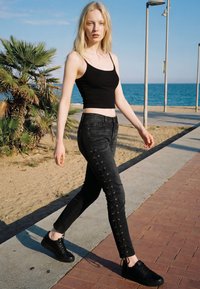 Tânără în maiou negru și blugi negri cu șireturi mergând pe faleza de lemn lângă plajă și ocean sub un cer senin.