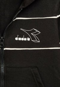 Giacca nera con zip realizzata in tessuto morbido, caratterizzata da strisce bianche e logo Diadora sul petto sinistro. Include una tasca frontale.