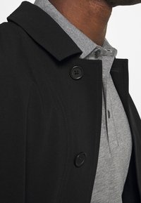 Svart kappa med spetsig krage och två svarta knappar, lagd över en grå pikétröja med en texturerad tyg och knäppning.