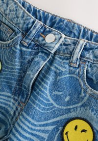Jean en denim bleu avec un bouton argenté, des motifs de visages souriants délavés et un patch smiley jaune qui fait un clin d’œil près de la poche.