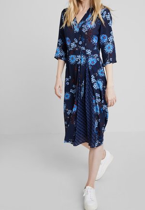 Kvinde iført en marineblå kjole med blå blomsterprint og hvide sneakers, der går mod en ensartet lys grå baggrund.