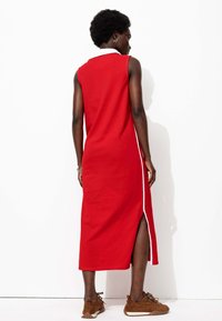 Robe sans manches rouge avec un col, fente sur le côté et bord blanc. Le modèle porte des chaussures texturées marron. Tissu lisse, design longueur cheville.